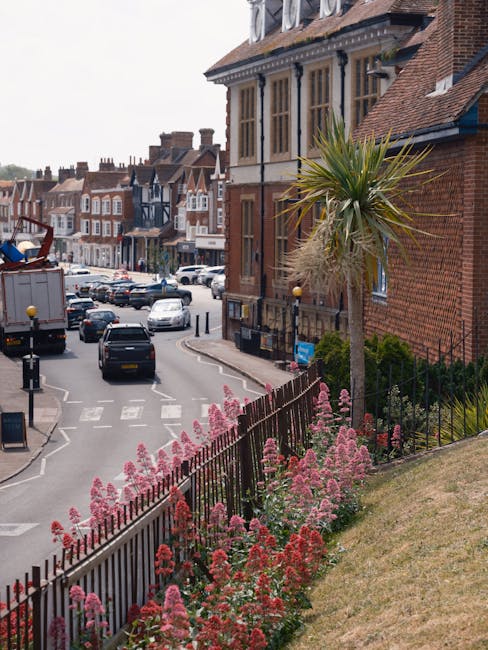 A street scene in Harpenden featuring a row of traditional brick buildings with bay windows and tiled roofs, along with a tall, slender palm tree and vibrant pink and red flowering plants along a blac