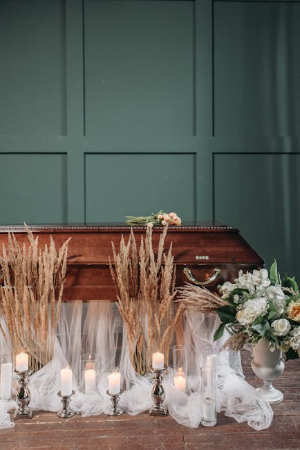 A floral arrangement displayed on a white, semi-transparent fabric-draped surface in front of a closed, green paneled wall. The arrangement includes white roses, peonies, and greenery in a white vase,