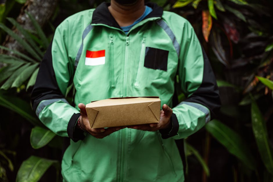 A delivery person wearing a bright green and black jacket with a Dutch flag patch on the left chest, holding a rectangular brown paper parcel with folded corners, standing outdoors among lush green fo