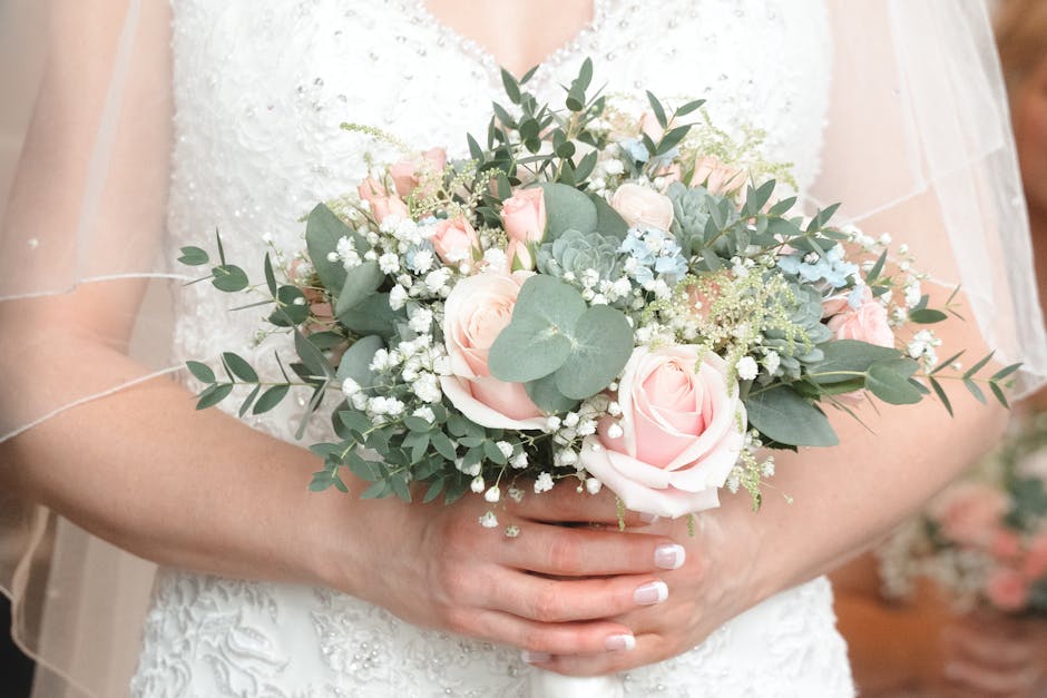 A bride holding a delicate floral bouquet featuring soft pink roses, white baby's breath, and eucalyptus leaves, arranged in a rounded, lush style. The bouquet is wrapped with a white ribbon, and the 