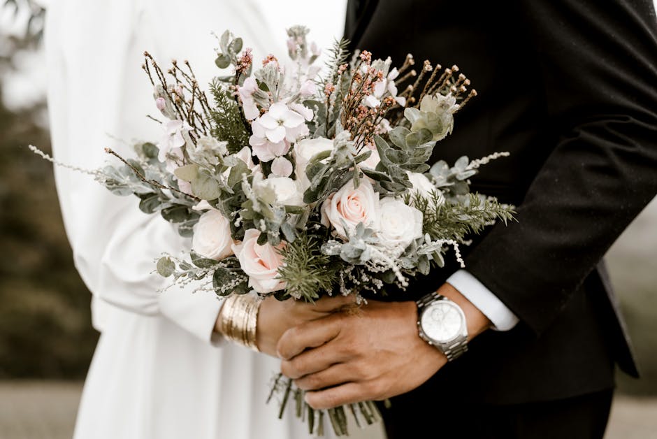 Close-up of a floral bouquet held by a bride and groom, featuring soft pink roses, white ranunculus, and delicate sprigs of eucalyptus and other greenery. The bouquet is finished with light pink and w